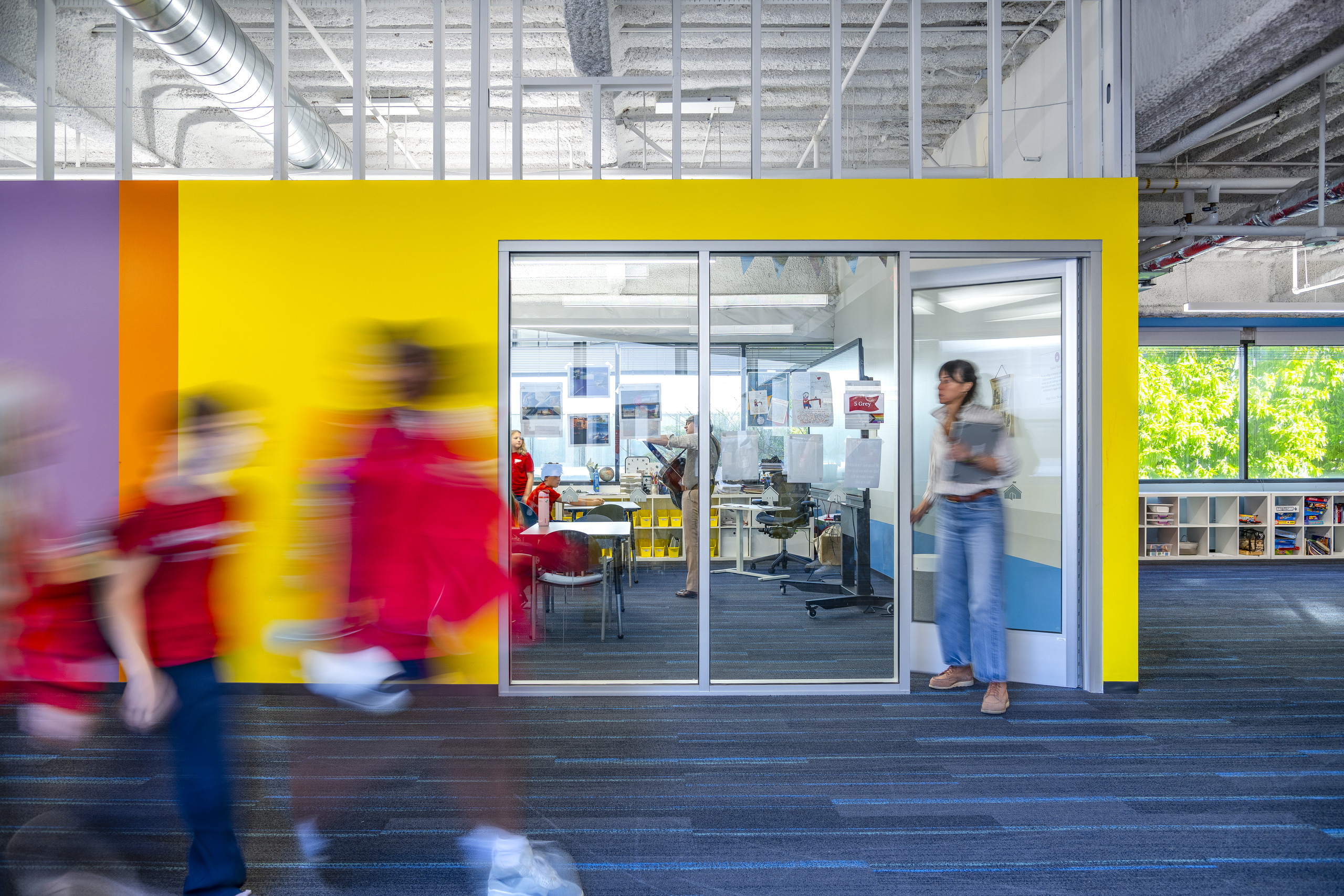 Students walking past a glass-enclosed classroom with a bright yellow wall at Village School in Santa Monica.